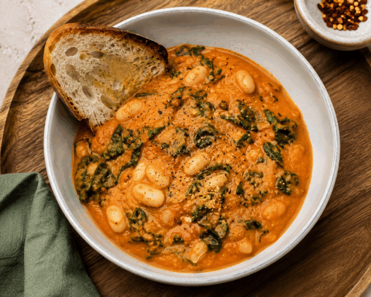 Creamy roasted tomato and cannellini bean stew with kale, served with toasted crusty bread in a ceramic bowl.