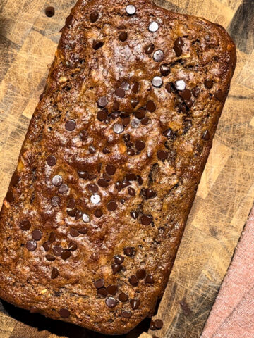 Chocolate chip high-protein banana bread baked in a loaf pan on a wooden butcher block, topped with mini chocolate chips and photographed from above. The loaf has a golden-brown crust with a moist texture, surrounded by scattered chocolate chips and a soft pink kitchen towel, creating a cozy homemade baking scene.