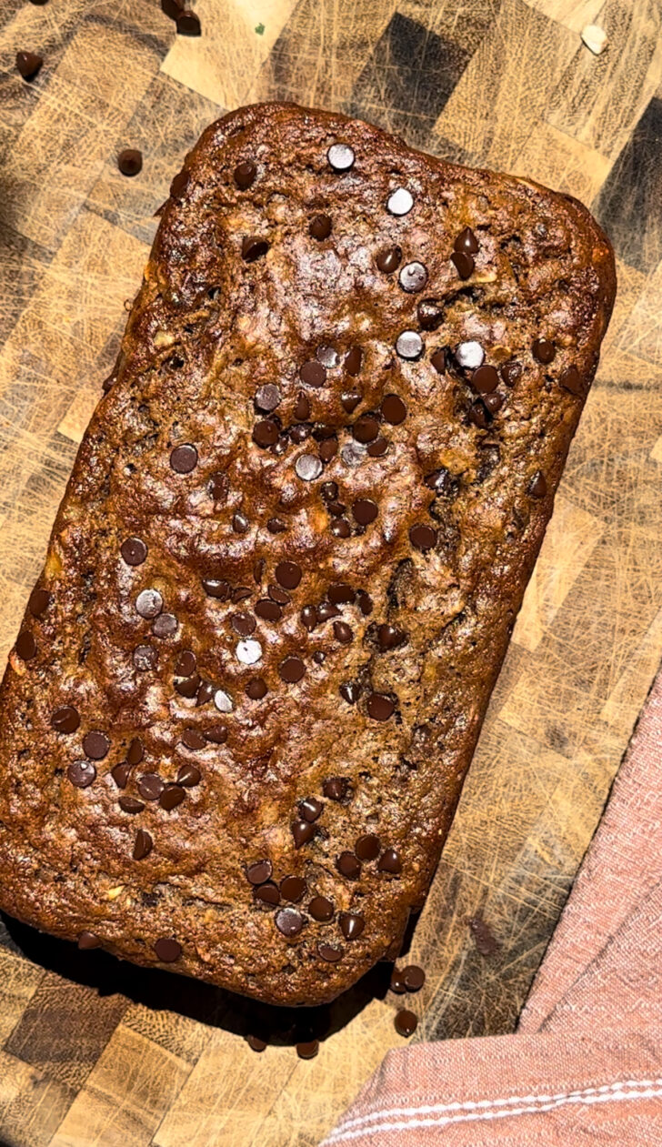 Chocolate chip high-protein banana bread baked in a loaf pan on a wooden butcher block, topped with mini chocolate chips and photographed from above. The loaf has a golden-brown crust with a moist texture, surrounded by scattered chocolate chips and a soft pink kitchen towel, creating a cozy homemade baking scene.