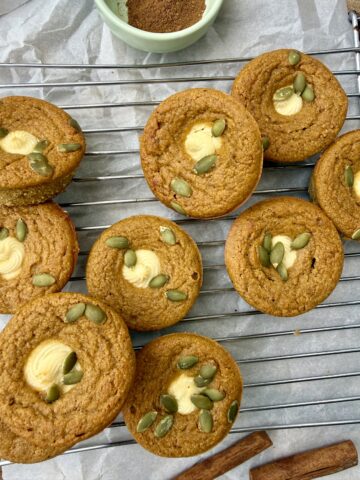 High-protein pumpkin cream cheese muffins topped with pepitas, cooling on a wire rack with cinnamon sticks and pumpkin spice for a cozy fall setting.