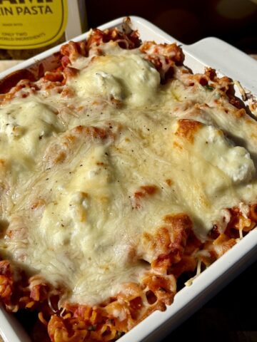 Golden, bubbly creamy tomato baked radiatori pasta topped with melted mozzarella, ricotta, and Parmesan cheese in a white baking dish, with Brami protein pasta packaging in the background.