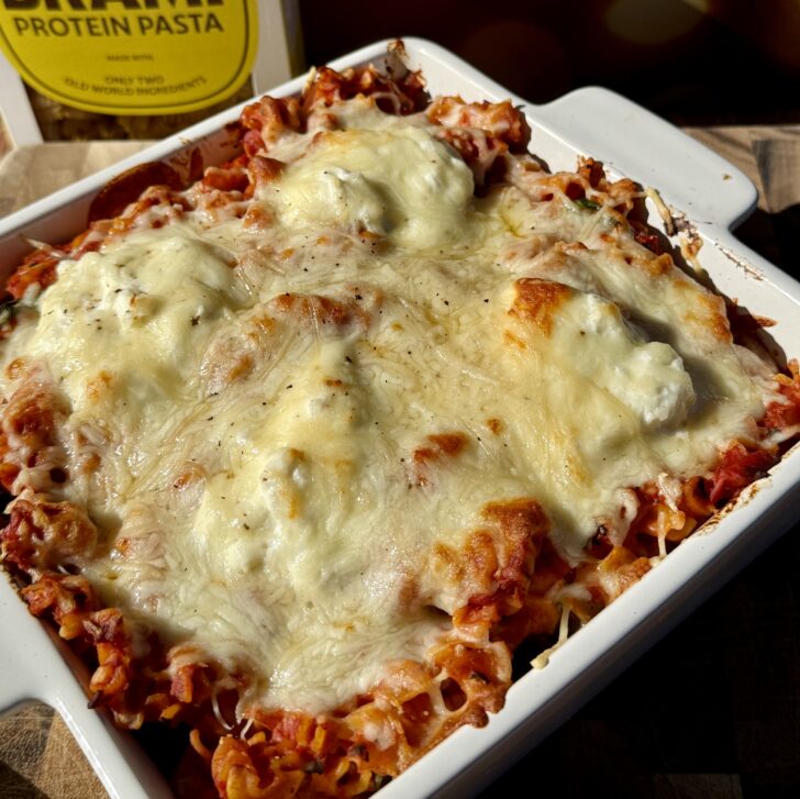 Golden, bubbly creamy tomato baked radiatori pasta topped with melted mozzarella, ricotta, and Parmesan cheese in a white baking dish, with Brami protein pasta packaging in the background.